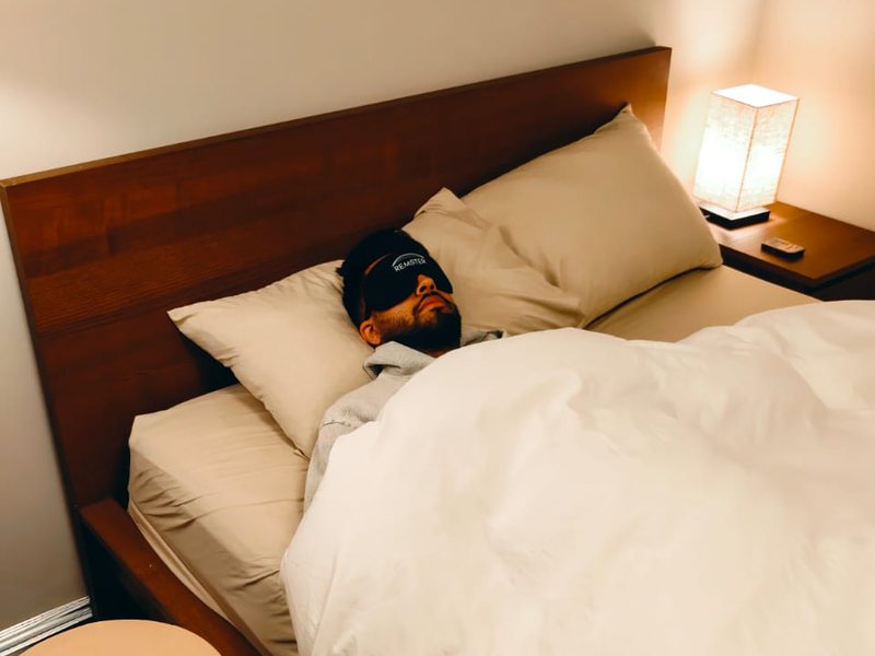 A man asleep in a bed wearing a Remster sleep mask, a walnut Remster nightstand beside the bed